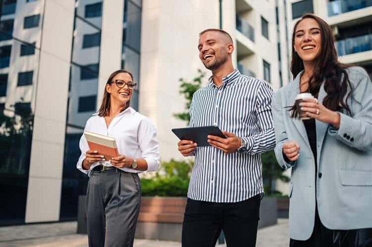Kolme iloista nuorehkoa ihmistä kävelevät kerrostalojen edessä. Yhdellä on kädessä kahvikuppi, toisella tabletti ja kolmannella kirja.